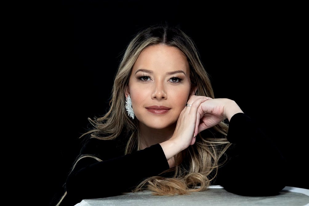 Retrato de la Dra. María Alejandra Ruiz, con las manos entrelazadas bajo el mentón, sobre fondo negro.
